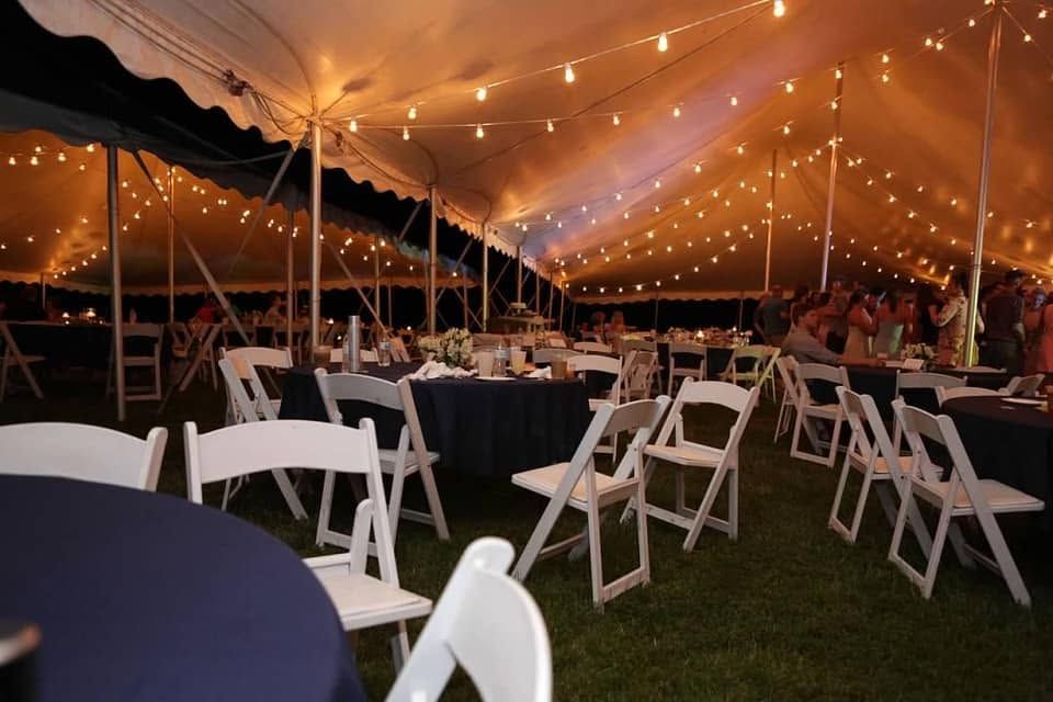 Evening wedding reception under a pole tent with string lights, navy linens and white chairs